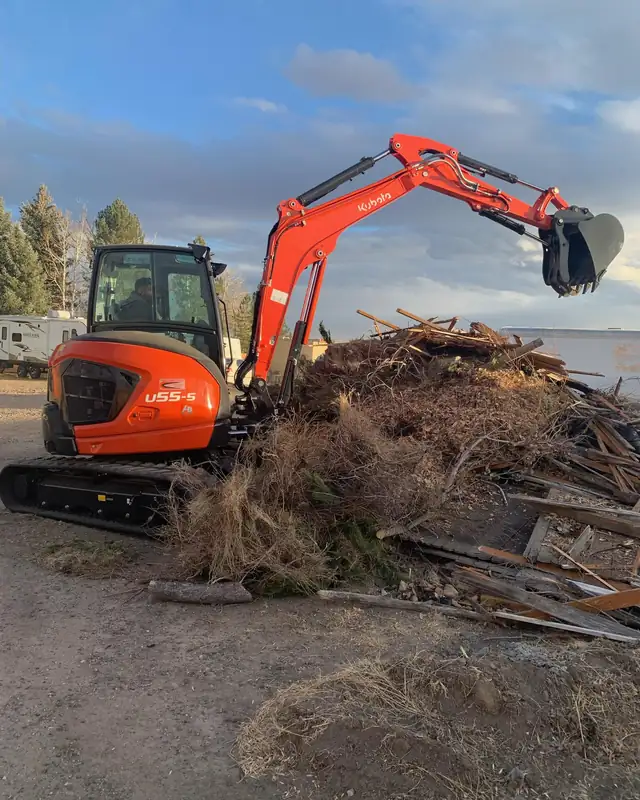 Orange mini excavator clearing a pile of old wood and brush from a yard