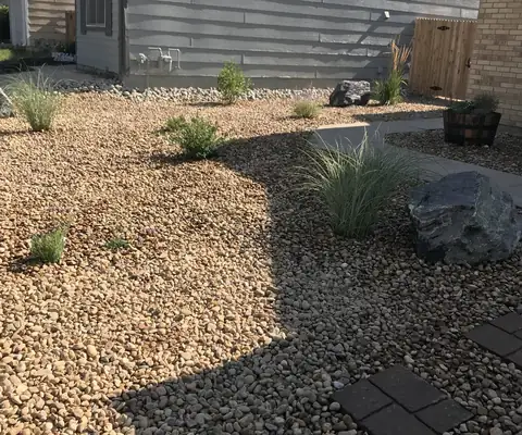 Side yard covered in rock with ornamental grasses, a boulder and square stepping stones